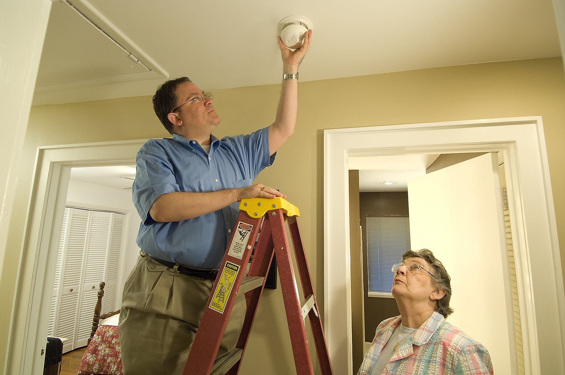 A person fitting a ceiling smoke alarm from a stepladder